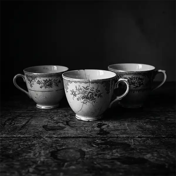 te para 3. Tres tazas de té antiguas sobre una mesa rústica de madera envueltas en sombras profundas con estilo dramático.