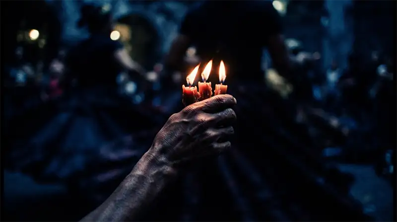 Primer plano cinematográfico de una mano sosteniendo velas encendidas con faldas en movimiento difuminadas al fondo bajo una luz cálida y nocturna.