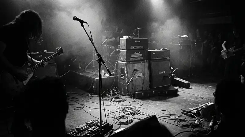 Escenario de rock ochentero lleno de cables y humo bajo luces tenues con estética analógica en blanco y negro.