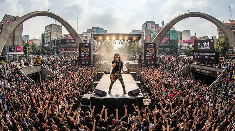 "Fotografía panorámica del concierto sorpresa de El Tri en la Glorieta de Insurgentes para celebrar los 50 años del Metro de la CDMX. Muestra a Alex Lora en un escenario central rodeado por una multitud de miles de personas que abarrotan la plaza y los accesos de la estación del Metro. Se aprecian los edificios circundantes y la estructura icónica de la Glorieta bajo la luz del día."