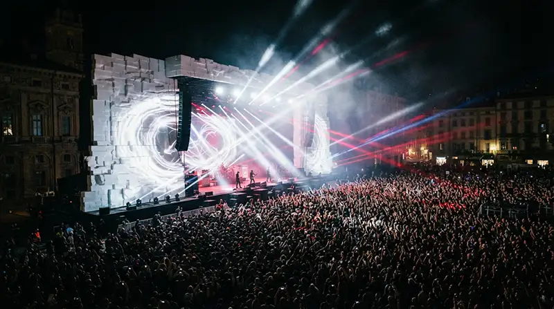 Fotografía épica de un concierto nocturno masivo frente a un gigantesco muro blanco iluminado por reflectores y láseres.