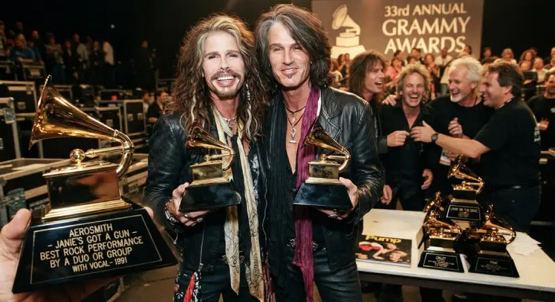 Steven Tyler y Joe Perry sonrientes en la ceremonia de los Premios Grammy de 1991, sosteniendo estatuillas doradas, con una placa grabada que acredita el galardón por "Janie's Got a Gun" en la categoría de mejor interpretación de rock.