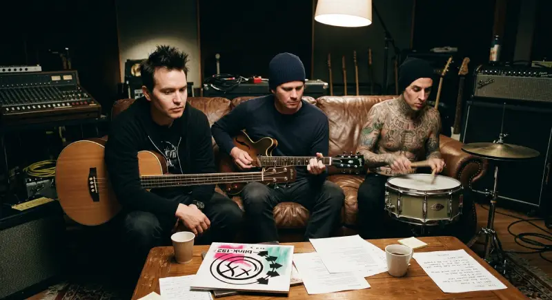 Una fotografía a color de Mark Hoppus, Tom DeLonge y Travis Barker, sentados en un sofá de cuero marrón en un estudio de grabación con poca luz. Mark Hoppus tiene cabello oscuro y pinchos, y sostiene un bajo acústico. Tom DeLonge usa un gorro y toca una guitarra eléctrica. Travis Barker está sin camisa y tatuado, y toca un tambor de trampa. Sobre una mesa de café frente a ellos, hay una copia del álbum "Blink-182" de 2003 con su logotipo de cara sonriente rosa y blanco, junto con hojas de letras dispersas y tazas de café.