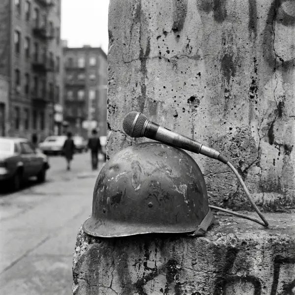 Fotografía en blanco y negro de un micrófono de estudio colocado sobre un viejo casco militar contra una pared de concreto, representando la intersección entre la música de protesta y el conflicto bélico.